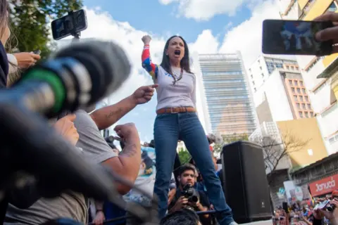 Jonathan Lanza/NurPhoto via Getty Images Venezuelan opposition leader Maria Corina Machado addresses a protest rally, raising her hands in the air as she speaks.