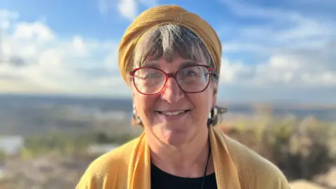 Standing outside, Sondra, wearing red-rimmed glasses, a yellow scarf, and a yellow cardigan, stares into the camera. Out of focus green in the background