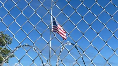 BBC An American flag flies against a blue sky behind a chainlink fence and barbed wire at Guantanamo Bay.