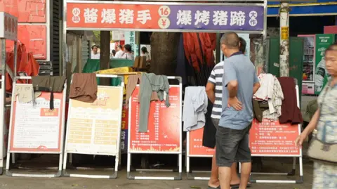 Xiqing Wang/ BBC A man in a t-shirt and shorts looks a row of red and yellow bulletin boards with job ads on them and clothes draped over them.