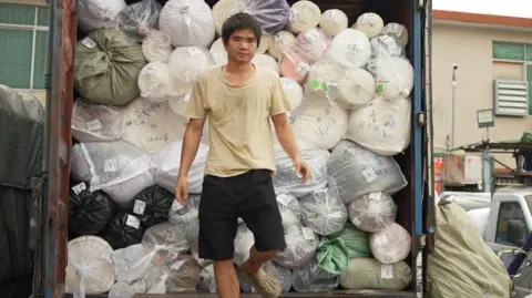 Xiqing Wang/ BBC A man in a beige t-shirt and black shorts is stepping down from a truck piled high with rolls of fabric of various colours - pink, white, green. 