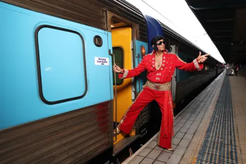 Don Arnold/WireImage An Elvis impersonator in a red jumpsuit points to the sky as he boards a train.