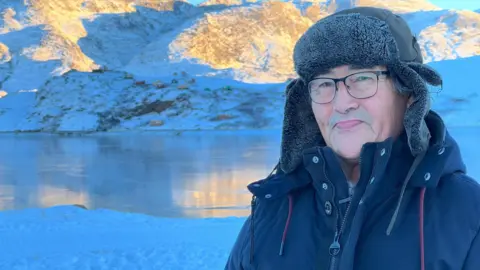 Kaaleeraq in a black jacket and hat smiling in front of some fish with a fjord in the Ringstead background