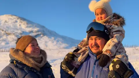 Angutimarik Hansen wearing goggles and a beanie holds a small child on his shoulder as his wife stands beside him and smiles.