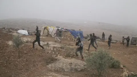 About a dozen B'Tselem men dressed in black, faces covered by hoods and scarves run in the same direction across dry ground, a small stone building in the background, Khirbet Susia in the southern Hebron hills, 21 December 2024.