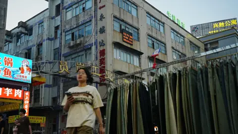 Xiqing Wang/ BBC A women wearing a t-shirt and shorts walks past a row of jeans on display on the street in the late evening. Behind her are buildings which house 
the factories.