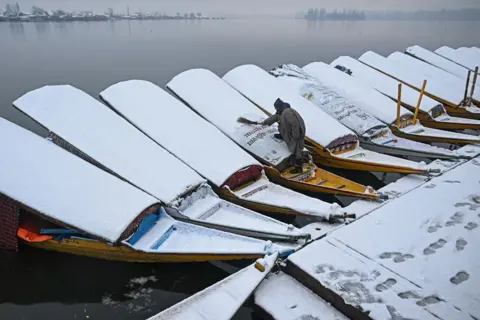 Tausif Mustafa/AFP Rows of boats covered in ice on a misty lake in India. A man in gray uses a brush to remove snow