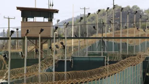 Reuters aerial photo shows high barbed wire fences, several cameras on poles and a watch tower at the Guantanamo Bay prison. In the background are trees and brush as well as another fence