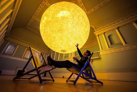 Ben Birchall/PA Luke Jerram sits on a chair and reaches out to his latest astronomical sculpture, Helios.