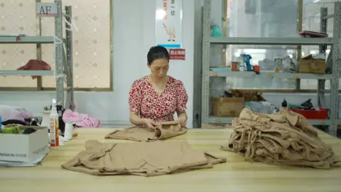 Xiqing Wang/BBC A woman in a red dress stands at a large wooden table folding brown t-shirts. A oile of folded brown trousers also sits on the table. 