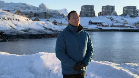 Kuno Fenker stands by the water in a blue jacket with the sun shining on his face