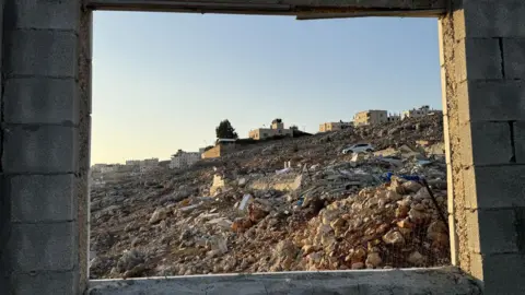 View through empty window frames of a large pile of rubble in Nablus, with some buildings still standing in the distance