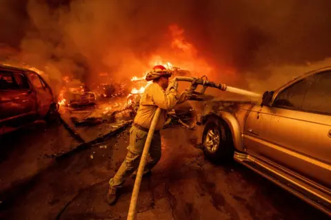 Ethane soap/AP flames can be seen in the background as a firefighter raises a water hose to spray a burning vehicle.