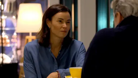 Malina Abelsen sits at a table wearing a blue shirt with a yellow cup in front of her