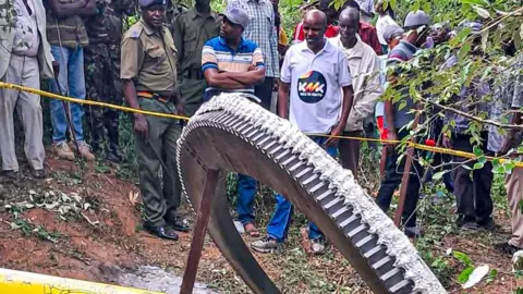 Getty Images A crowd, including police officers, stands behind yellow tape as they watch a giant metal ring fall from space onto farmland in Mukuku, Kenya. 