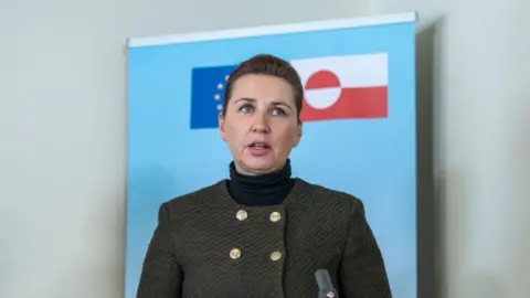 Getty Images Danish Prime Minister Mette Frederiksen, wearing a dark green textured blazer with gold buttons and a black turtleneck, speaks at a press event. He stands in front of a light blue background featuring the flags of Greenland and the European Union. His expression is serious as he addresses the audience with a microphone visible in the foreground.