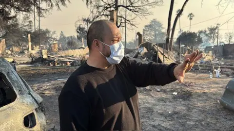 Larry Villescas stands in front of the charred remains of his neighborhood. 